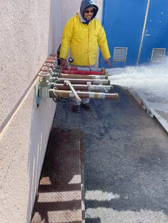 An IFS technician in a yellow rain jacket flow testing a bank of standpipe outlets, water discharging across the pavement