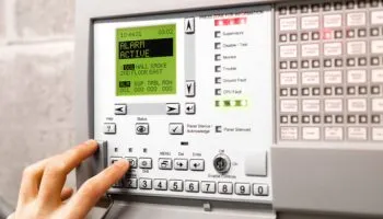 A hand pressing the keypad of a fire alarm control panel, its display reading ALERT ACTIVE beside a column of lit zone status indicators