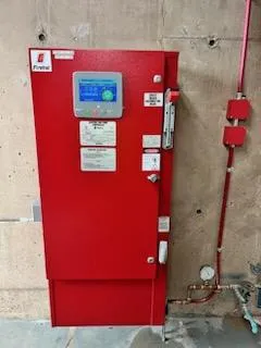 Red Firetrol fire pump controller cabinet wall-mounted on concrete, with a digital status display and sensing line running to the riser