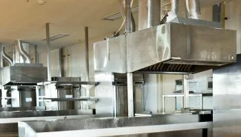 Rows of stainless steel exhaust hoods above a commercial kitchen cooking line, with suppression nozzles and ductwork running to the ceiling