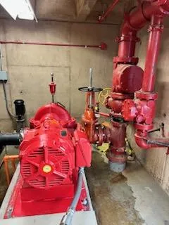 Red electric fire pump and motor in a pump room, connected to red riser piping, control valves, and a pressure gauge manifold
